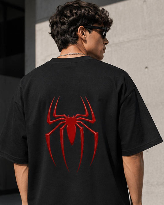 A man wearing a black oversized t-shirt with a large red spider graphic on the back, photographed from behind against a concrete wall.