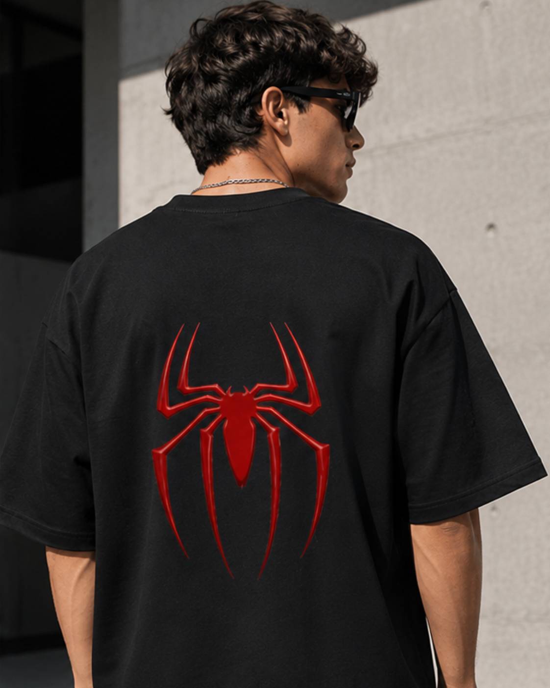 A man wearing a black oversized t-shirt with a large red spider graphic on the back, photographed from behind against a concrete wall.